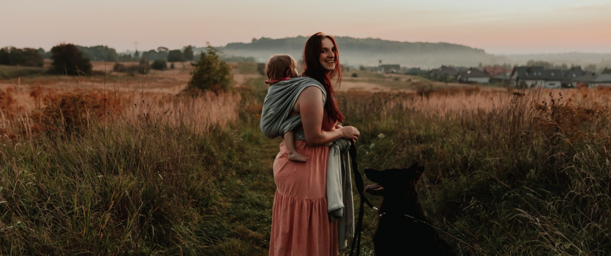 Matka stojąca na łące z córką w chuście na plecach obok, której stoi owczarek niemiecki.
